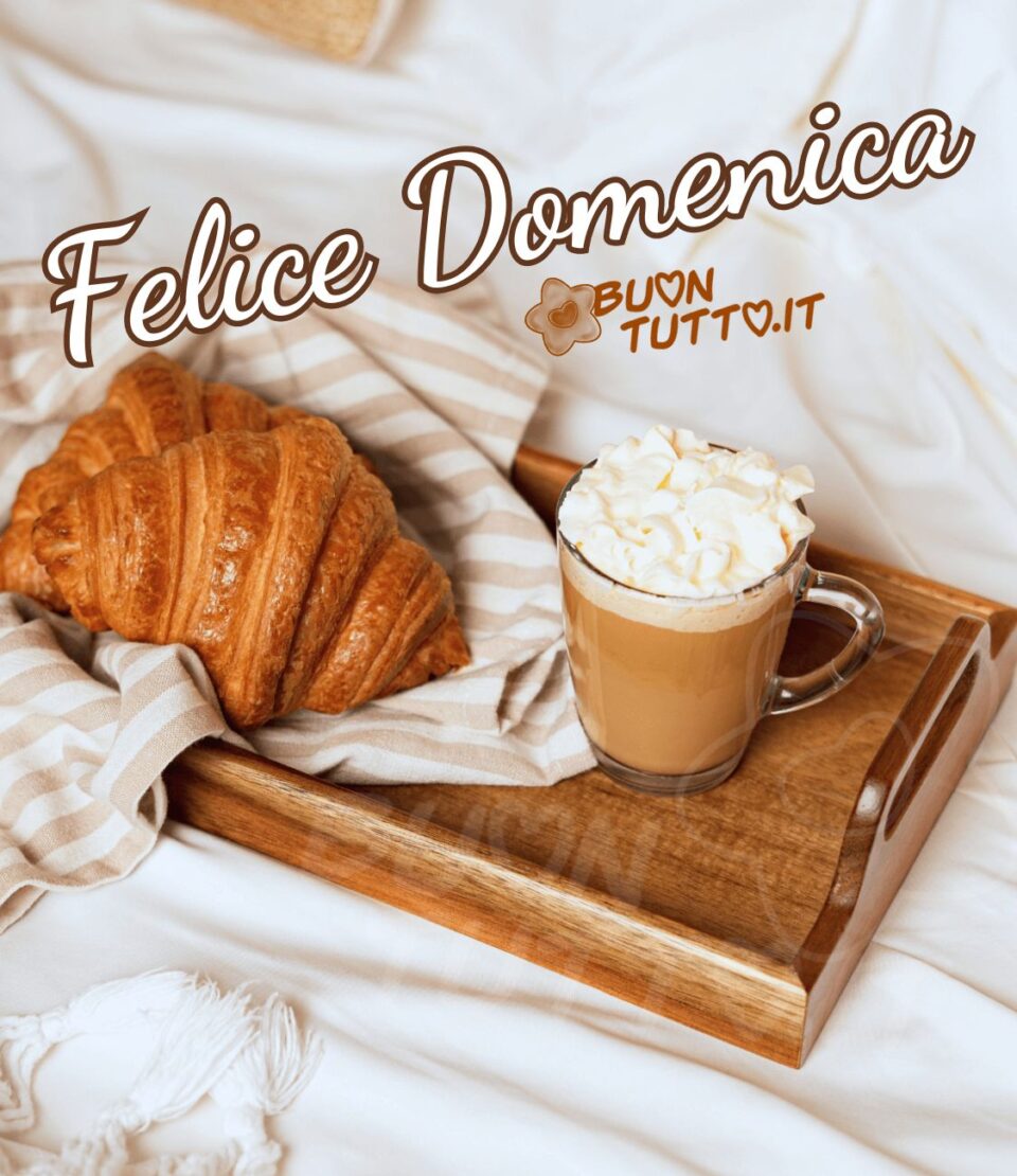 Foto di un vassoio di legno appoggiato sopra un letto con sopra un tovagliolo di cotone con righe orizzontali bianche e nocciola con sopra due fragranti cornetti e una gustosa tazza di vetro di caffellatte con panna montata. Nella parte alta dell'immagine c'è scritto in un carattere corsivo elegante felice domenica di colore bianco e contorno marroncino. Una bellissima immagine che trasmette relax e buonumore da scaricare gratis e condividere con amici parenti gruppi tramite WhatsApp Facebook Twitter Pinterest Telegram Instagram autore buontutto.it 