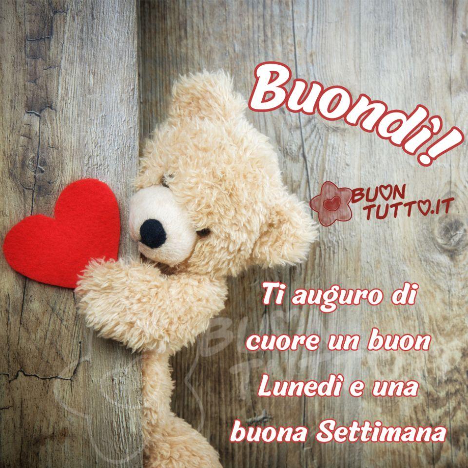 Foto di un orsetto di peluche, di colore cremino dal tenero musetto  , che si affaccia da un tronco d'albero tenendo nella zampina un grande cuore rosso. Nella parte alta a destra dell'immagine, vicino la testa dell'orso, c'è scritto in forma semi-circolare di colore bianco e contorno bordeaux Buondì!; Nella parte bassa a destra c'è scritto di colore bianco e contorno rosso sfumato Ti auguro di cuore un buon lunedì e una buona settimana. Un'immagine che trasmette tenerezza, affetto e serenità da scaricare gratis e condividere con amici, parenti, gruppi e su tutte le tue piattaforme social preferite creata da buontutto.it