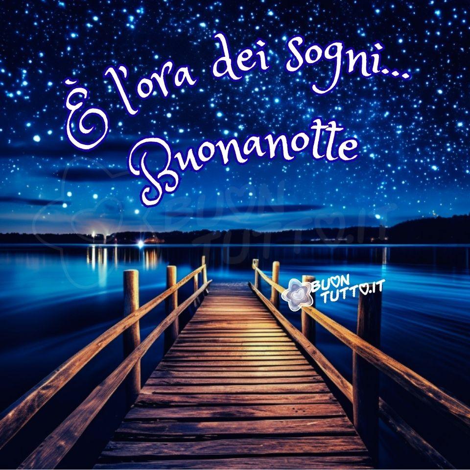 Foto di un molo di legno che si estende sul mare calmo che sembra uno specchio d’acqua. La prospettiva è dal molo, guardando dritto verso l'orizzonte. Il molo è fatto di assi dalle venature marroni chiare e scure e ha una semplice ringhiera di legno su entrambi i lati. All’orizzonte si intravede un panorama fato di montagne e colline e alcune luci che possono far pensare ad un paese. Il cielo è notturno, di un blu profondo intenso e vivace, costellato da una miriade di stelle luminose e scintillanti. Nella parte alta dell'immagine c'è scritto in forma semi-circolare in un carattere corsivo elegante con ghirigori di colore bianco e contorno azzurro È l’ora dei sogni… Buonanotte. Un'immagine romantica e bellissima che trasmette serenità, forza, pace, bellezza della natura, meraviglia e stupore da scaricare gratis e condividere con amici, cari, familiari, parenti gruppi tramite WhatsApp, Facebook, X Twitter, Pinterest, Telegram, LinkedIn, Instagram e le tue piattaforme social preferite. Autore buontutto.it