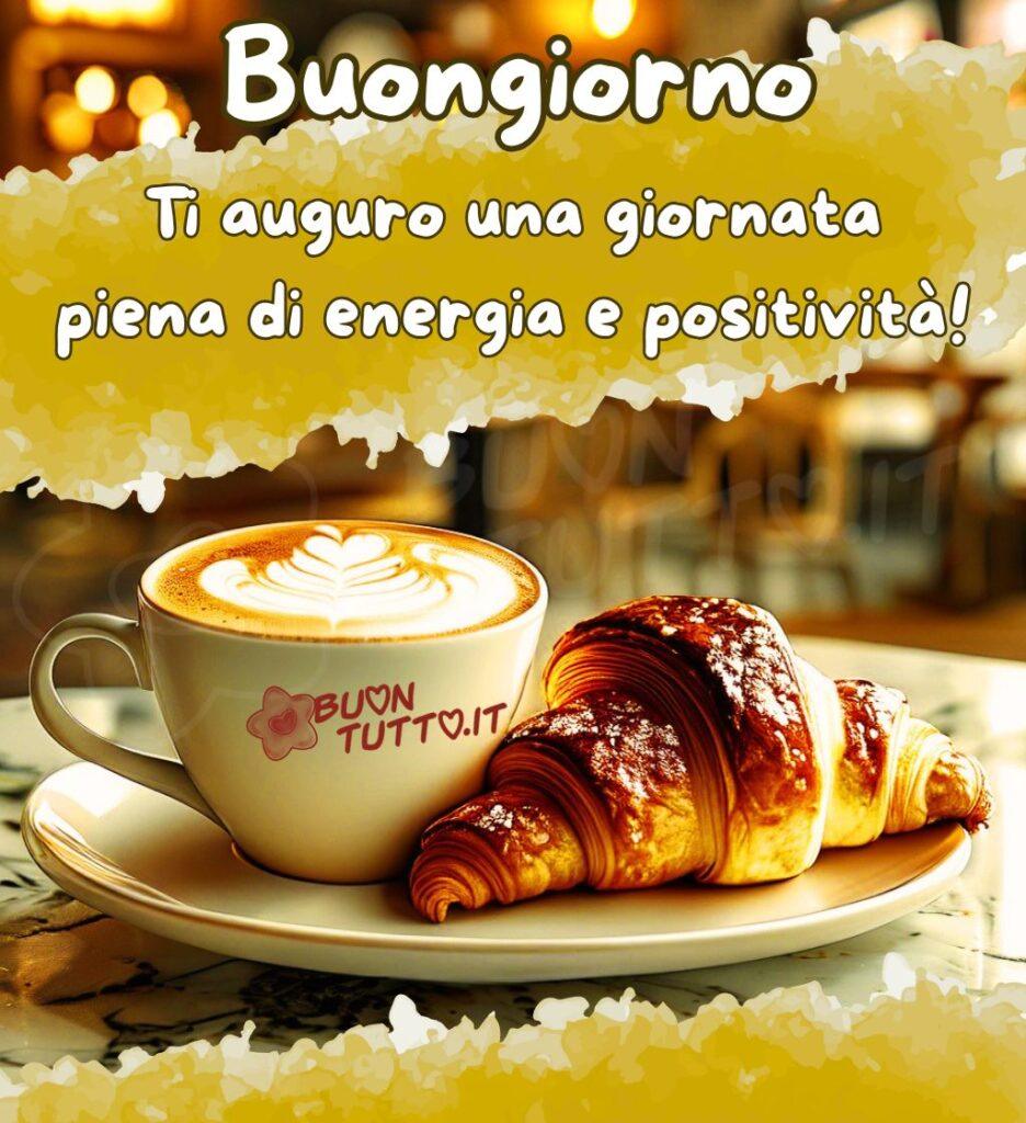 Un'immagine che ritrae su un piatto di ceramica una tazza di cappuccino dalla schiuma vellutata, decorata con un elegante disegno di latte art a foglia e un cuore, che irradia un calore invitante. Accanto, un croissant dalla sfoglia dorata e croccante, cosparso di un velo di zucchero a velo. Poggiati su un tavolo di marmo. Lo sfondo è una caffetteria immersa in una luce calda e soffusa (un meraviglioso effetto bokeh). L'atmosfera è resa ancora più accogliente e serena da un effetto artistico creato da due larghe pennellate di giallo-ocra brillante una in alto e una in basso, con bordi bianchi e frastagliati. Nella parte alta dell'immagine c'è scritto in un carattere corsivo bombato di colore bianco e contorno marrone: Buongiorno Ti auguro una giornata piena di energia e positività! È un'immagine che trasmette comfort, dolcezza, energia, accoglienza, bellezza, positività, serenità, tranquillità e pace da scaricare gratis e condividere con amici, cari, familiari, parenti gruppi tramite WhatsApp, Facebook, X Twitter, Pinterest, Telegram, LinkedIn, Instagram e le tue piattaforme social preferite. Autore BuonTutto.it