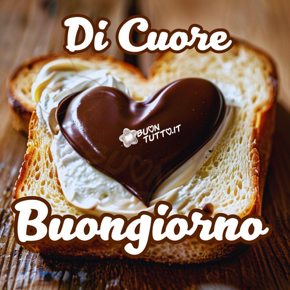 Foto in primo piano di una fetta di pane tostato in stile brioche, farcita in modo ordinato e appoggiata su una superficie di legno chiaro con venature visibili. Al centro della composizione spicca un grande cuore di cioccolato fuso. La superficie del cioccolato è estremamente lucida, quasi a specchio, e suggerisce una consistenza densa e vellutata, simile a una ganache o a una glassa a specchio. La fetta di pane, spessa e probabilmente di tipo brioche o pane in cassetta artigianale, è leggermente tostata, di forma rettangolare con bordi irregolari. Presenta una crosta ben dorata e una mollica soffice e alveolata; la sua forma richiama vagamente il profilo di un cuore. Sulla superficie del pane è steso uno strato generoso di crema bianca che potrebbe essere crema spalmabile, burro morbido, formaggio cremoso o panna montata distribuita con un movimento circolare che crea leggere onde. Al centro, sopra la crema, è posizionato un cioccolatino a forma di cuore, lucido e di colore marrone scuro, con superficie liscia e riflessi che indicano una consistenza compatta e brillante. La forma del cuore è ben definita e leggermente in rilievo rispetto alla crema. Nella parte alta dell’immagine compare la scritta Di Cuore in carattere corsivo bombato di colore bianco con contorno marrone; nella parte bassa, nello stesso stile, la parola Buongiorno. L’immagine trasmette dolcezza, accoglienza, cura ed eleganza, ed è ideale per rappresentare una colazione speciale o un gesto d’affetto da scaricare e condividere gratuitamente con amici, familiari, gruppi e contatti tramite WhatsApp, Facebook, X/Twitter, Pinterest, Telegram, LinkedIn, Instagram e altre piattaforme social. Autore BuonTutto.it